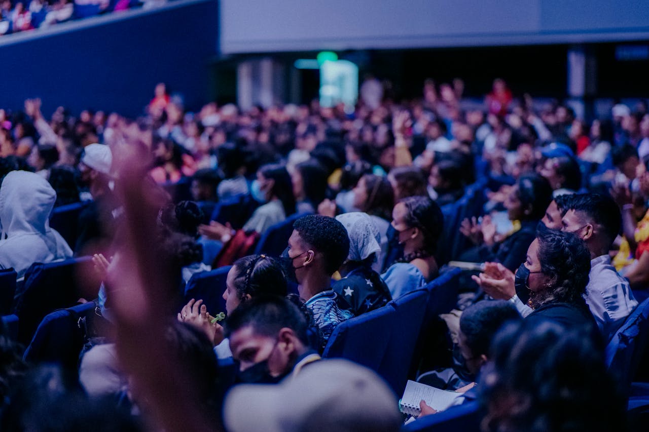 services-bg A large, diverse audience attentively watching a live event indoors, seated in rows.