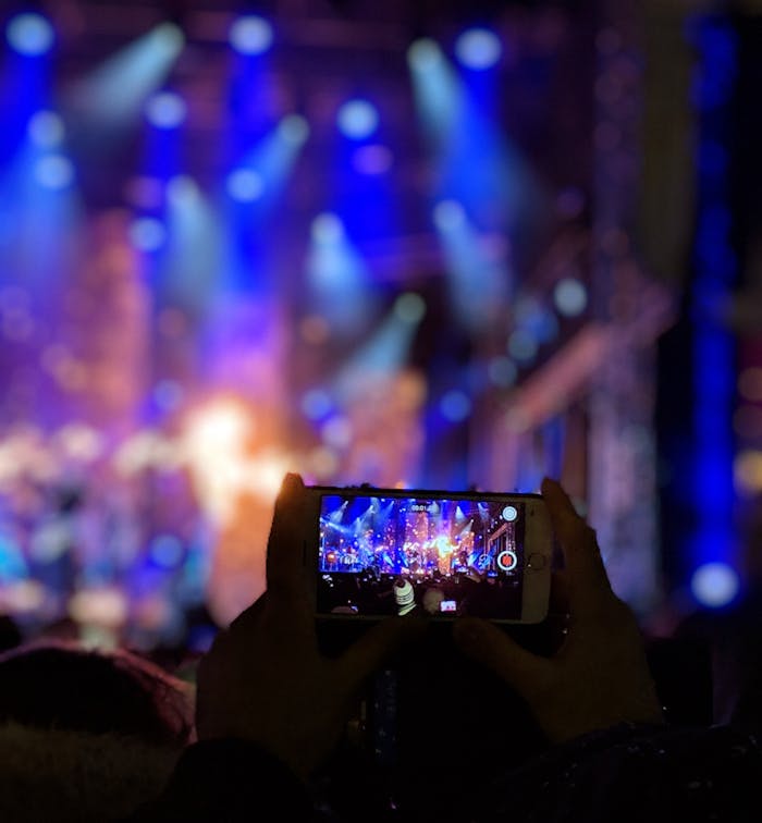our-story Silhouetted hands holding a smartphone capturing vibrant concert lights.